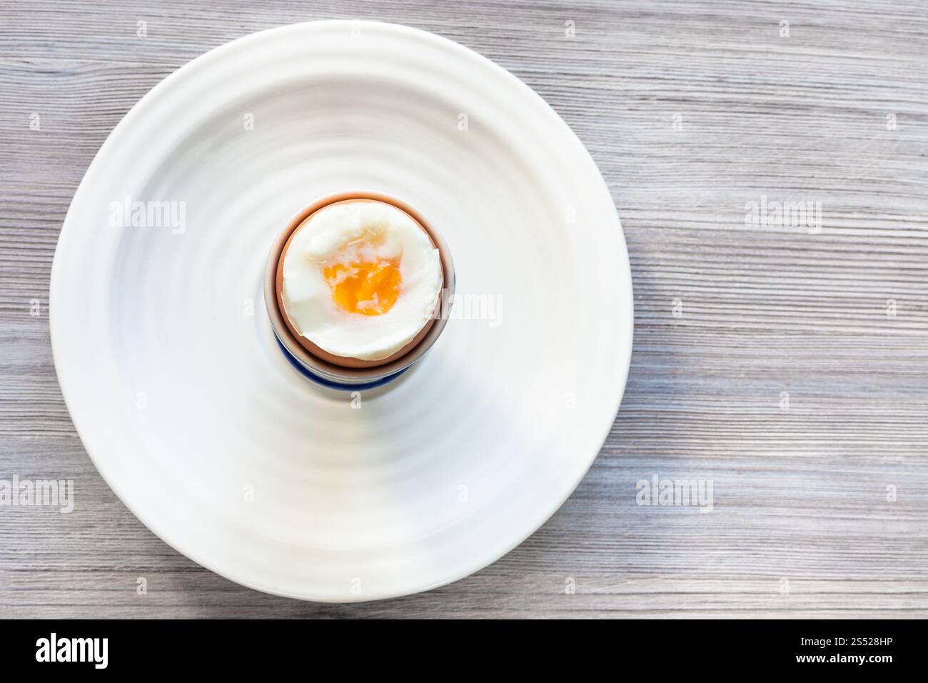 Vista dall'alto di soft-boiled brown uovo in tazza sulla piastra bianca su grigio asse di legno con copyspace vuoto Foto Stock