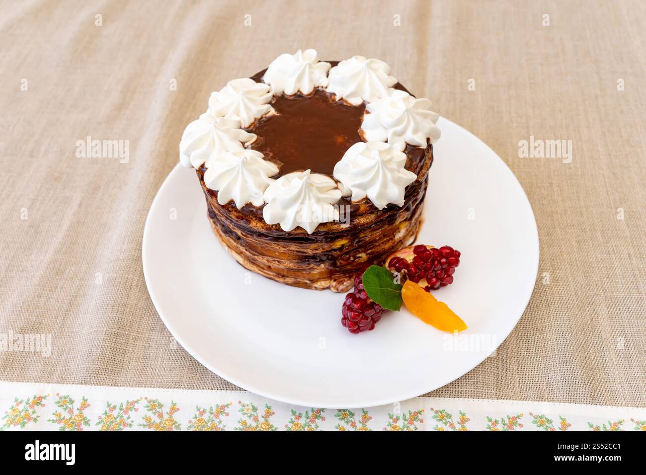 Una ricca torta al cioccolato adornata con panna montata si trova su un piatto bianco. Lamponi freschi e fette di arancia accompagnano il dessert su un tableclot beige Foto Stock