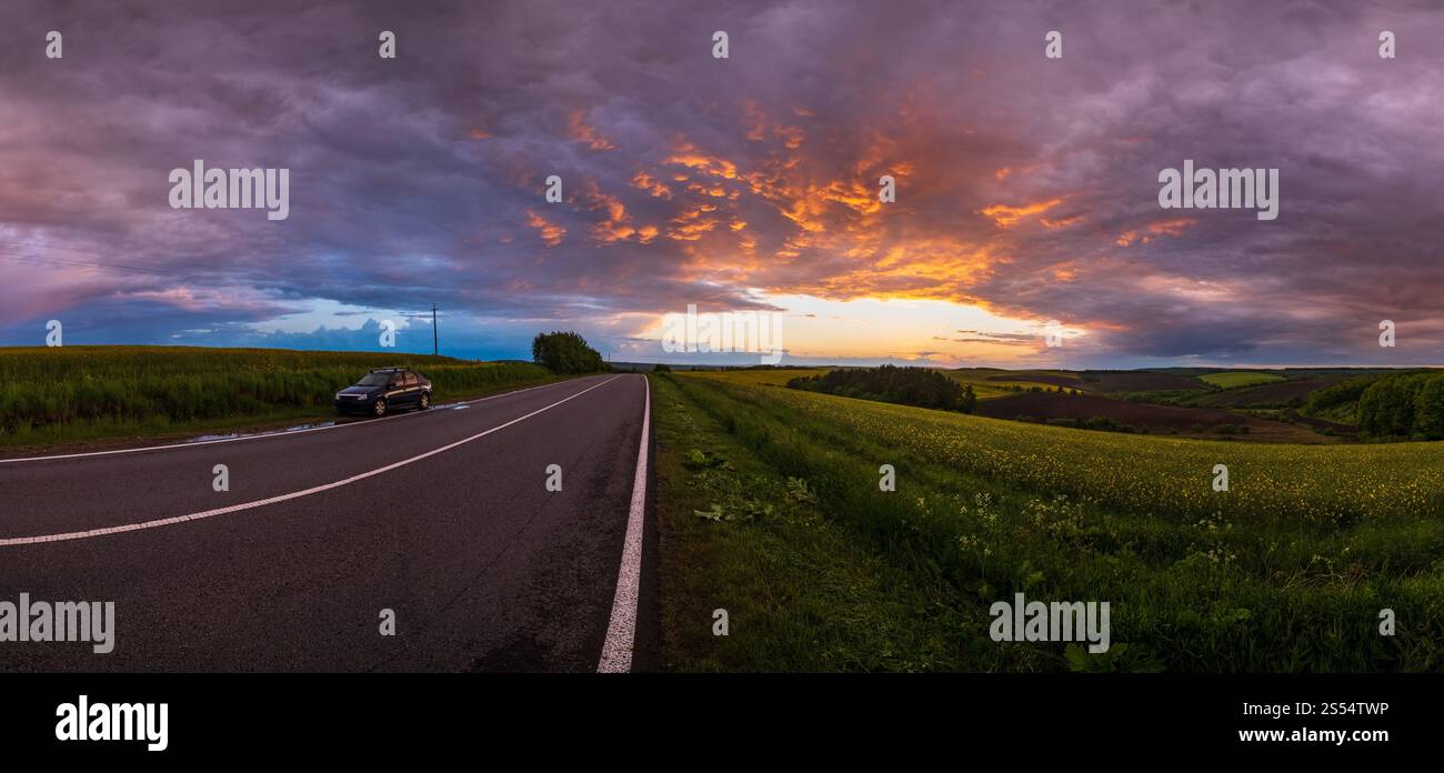 Campi gialli di colza primaverile, cielo notturno nuvoloso al tramonto, colline rurali e strada regionale. Stagione naturale, viaggi, clima, bellezza della campagna Foto Stock