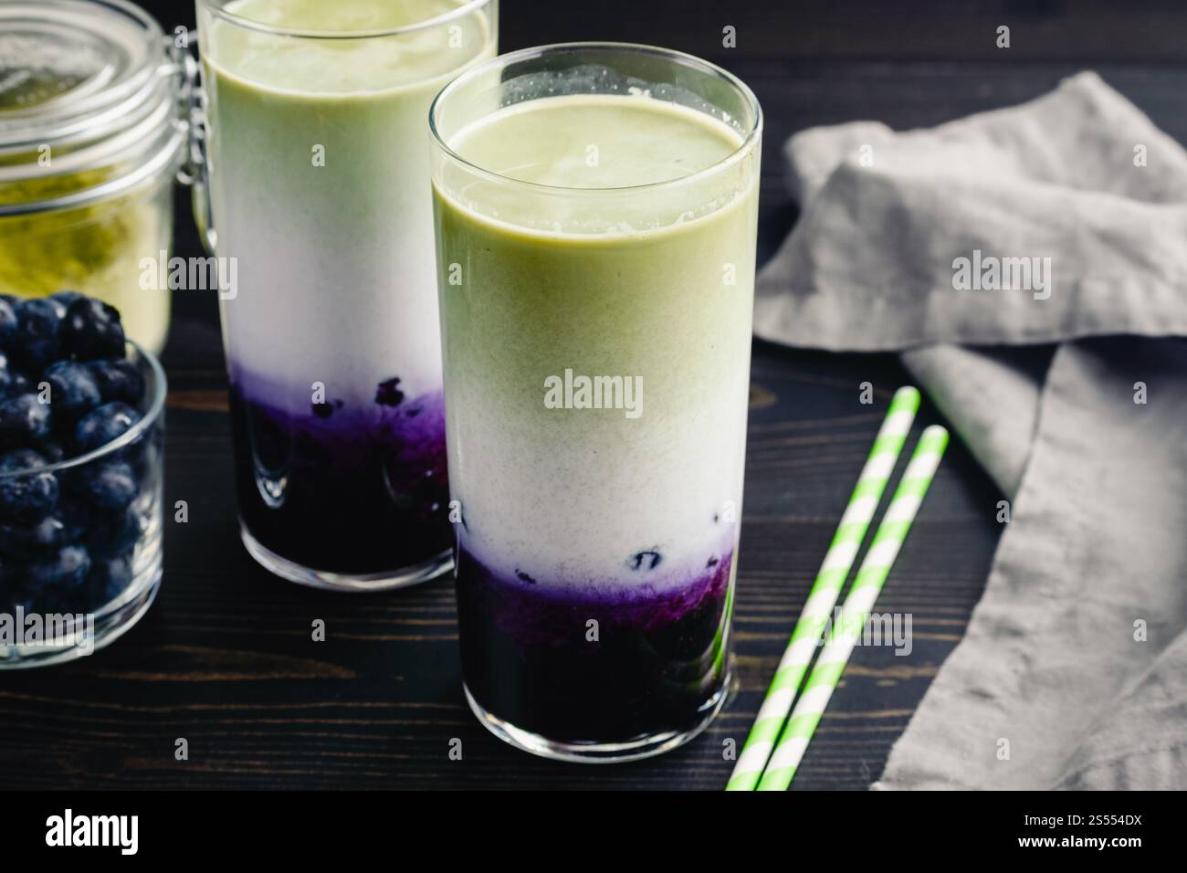 Lattine di Matcha al mirtillo ghiacciato in bicchieri alti: Lattine fredde a strati di tè verde, latte e mirtilli con cannucce di carta e ingredienti nelle vicinanze Foto Stock