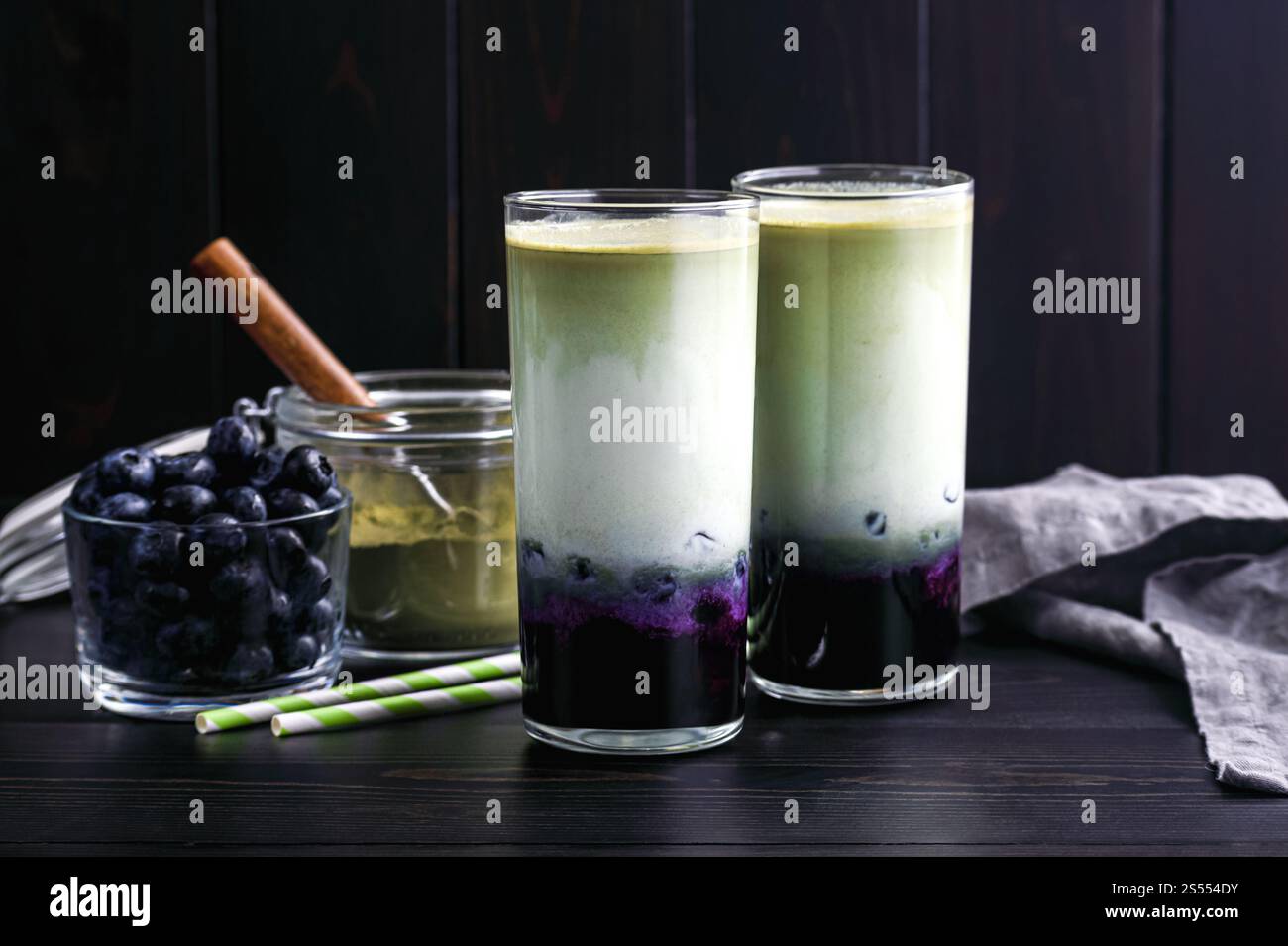 Lattine di Matcha al mirtillo ghiacciato in bicchieri alti: Lattine fredde a strati di tè verde, latte e mirtilli con cannucce di carta e ingredienti nelle vicinanze Foto Stock