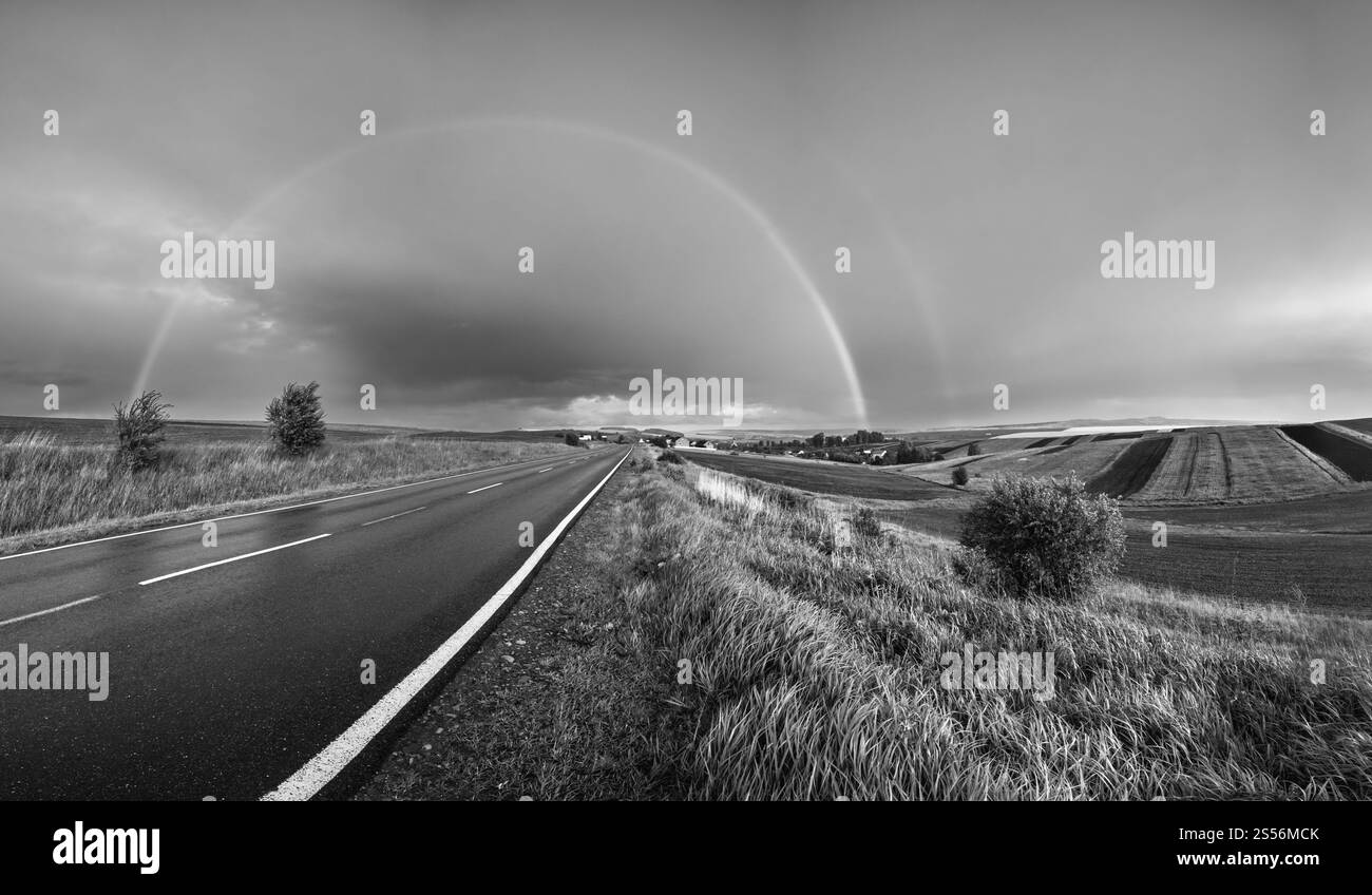 Scala di grigi. Colza primaverile e piccoli campi agricoli dopo la pioggia vista serale, nuvoloso cielo prima del tramonto con arcobaleno e colline rurali. Stagionale, meteo, Foto Stock