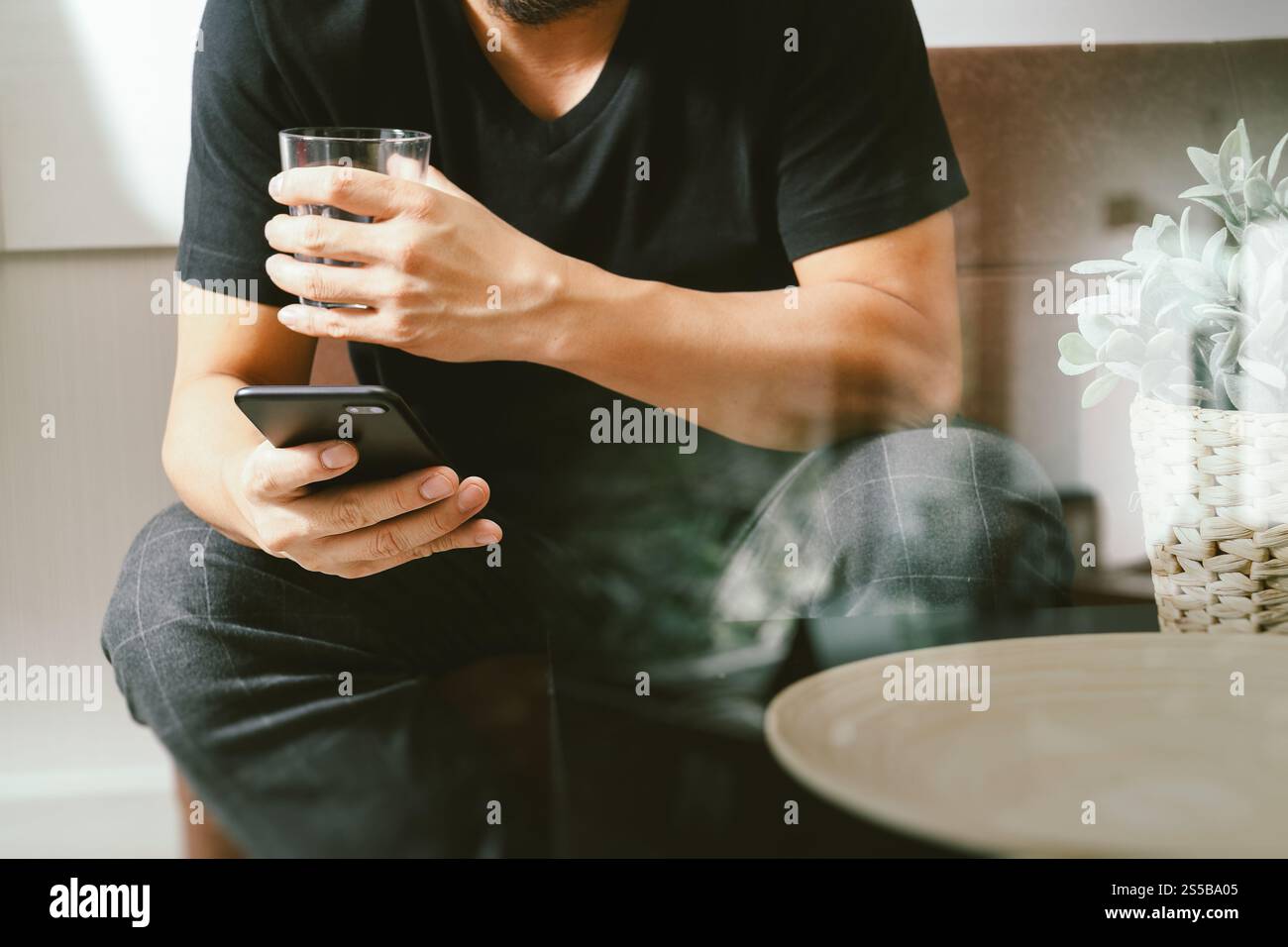 uomo di design a mano con un bicchiere d'acqua che usa il phon intelligente per i pagamenti mobili per lo shopping online, canale omni, seduto sul divano nel soggiorno, vaso in rattan Foto Stock