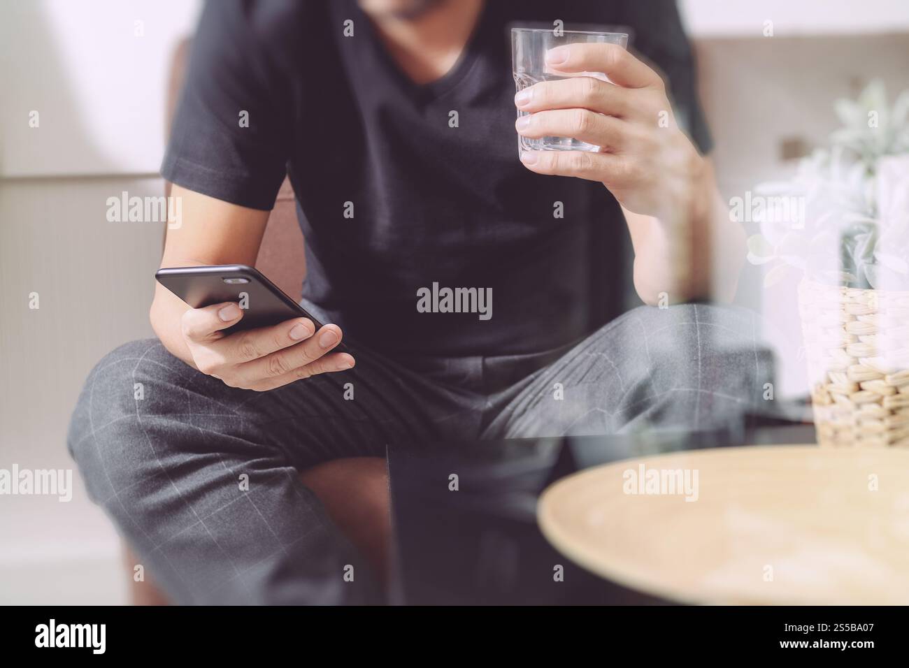 uomo di design a mano con un bicchiere d'acqua che usa il phon intelligente per i pagamenti mobili per lo shopping online, canale omni, seduto sul divano nel soggiorno, vaso in rattan Foto Stock