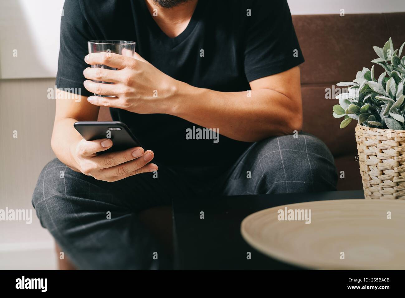 uomo di design a mano con un bicchiere d'acqua che usa il phon intelligente per i pagamenti mobili per lo shopping online, canale omni, seduto sul divano nel soggiorno, vaso in rattan Foto Stock