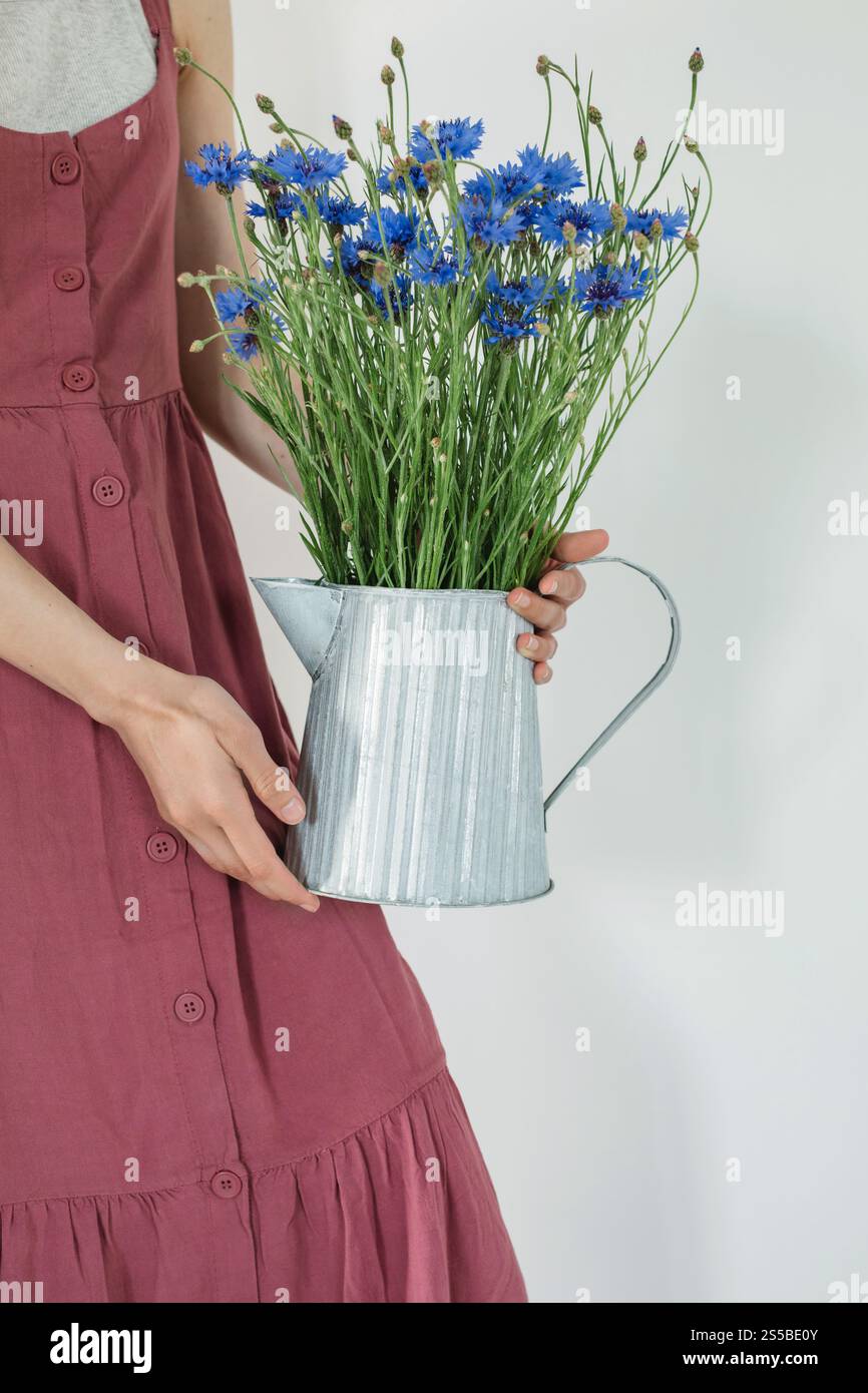 Primo piano di una donna in un prendisole estivo che tiene in mano una brocca metallica con un bouquet di fiori di mais blu Foto Stock