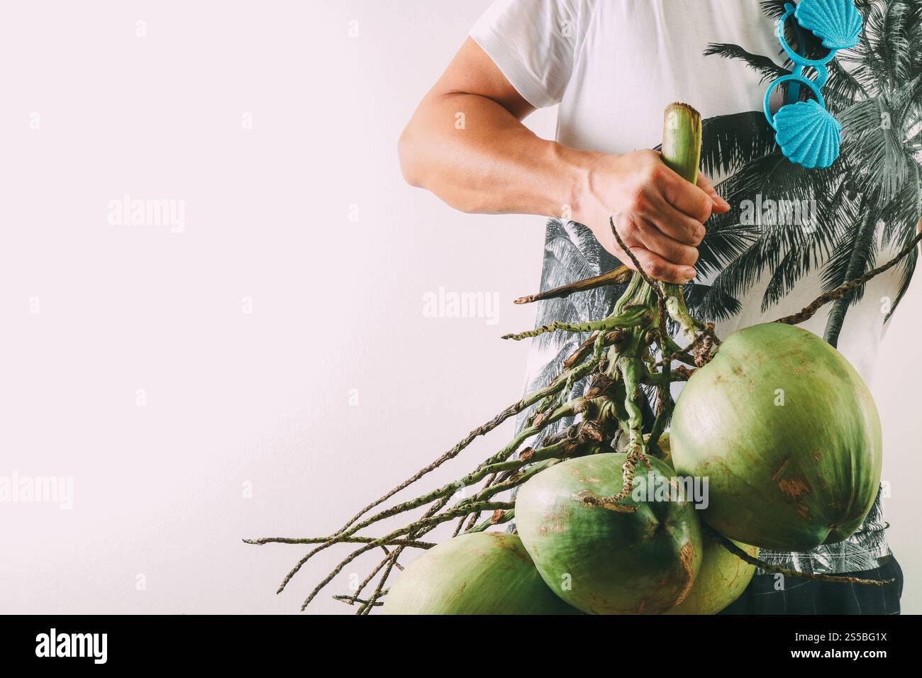 Concetto di moda per l'estate e le vacanze. Uomo che indossa le grafiche delle palme su una T-shirt e tiene un gruppo di noci di cocco con oggetti su sfondo bianco Foto Stock