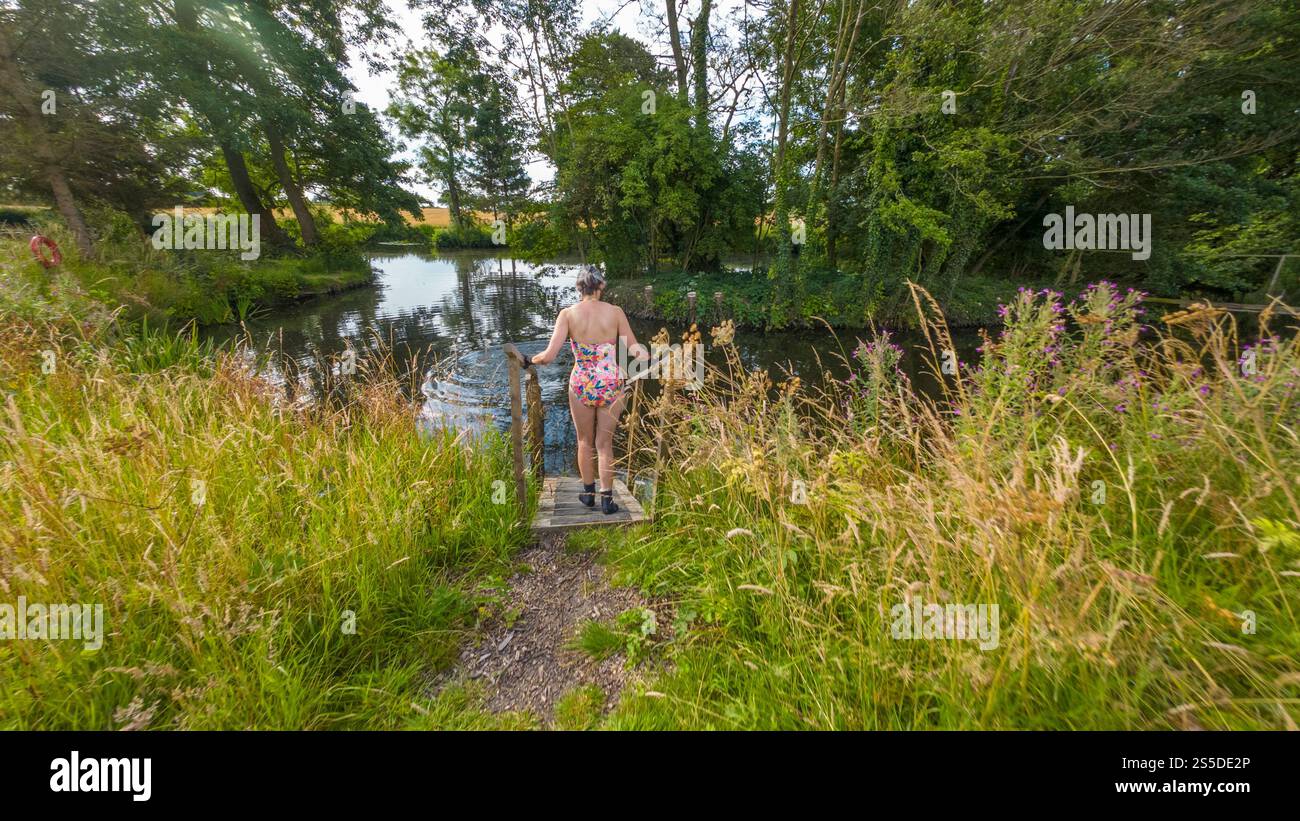 Vista posteriore di una nuotatrice selvaggia che indossa un costume da bagno dai colori vivaci, guanti in neoprene e stivali che entrano in uno stagno da nuoto con gradini di legno. Foto Stock