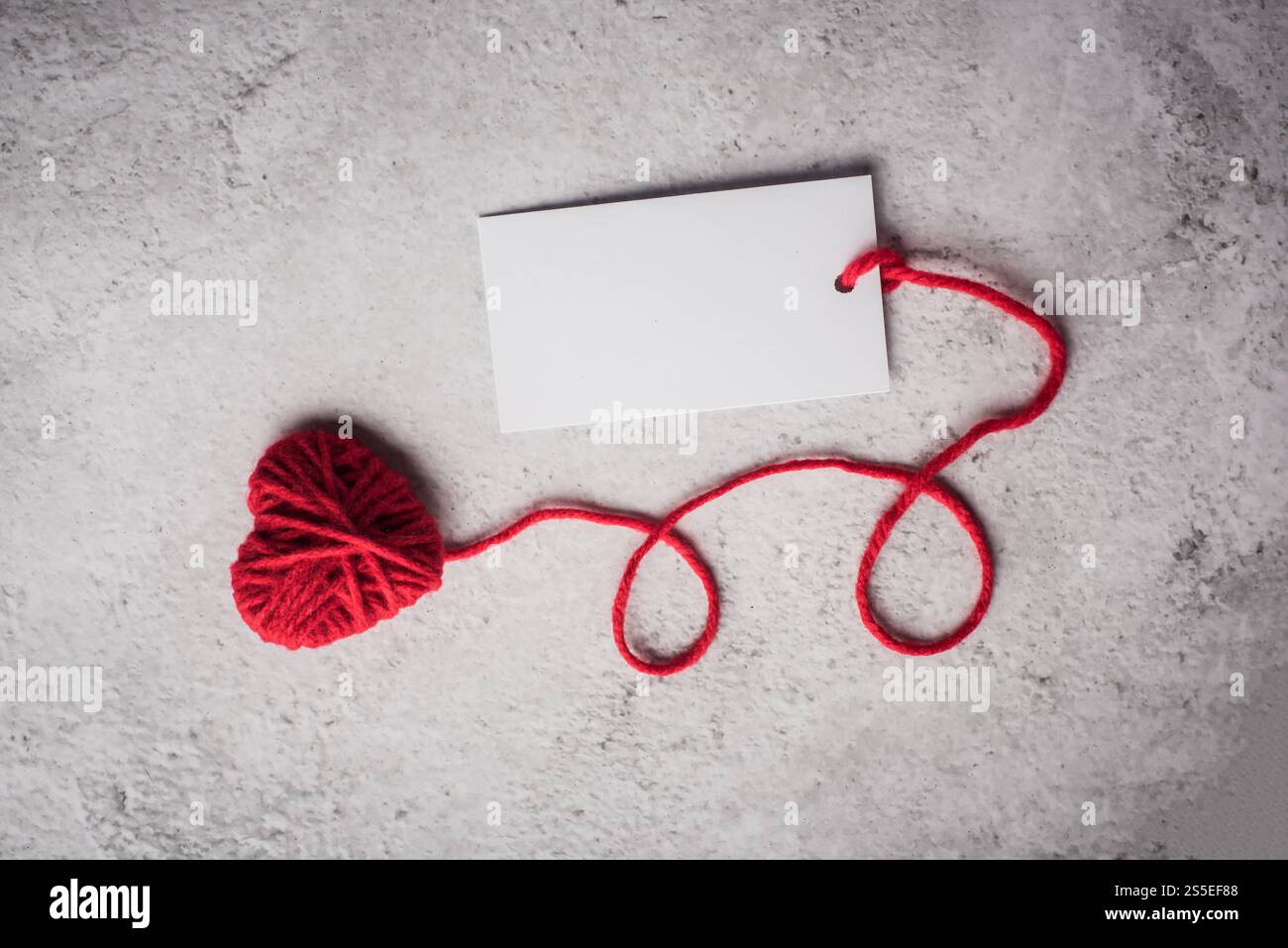 Cuore in filato rosso con cartellino bianco sullo sfondo della parete Foto Stock Cuore in filato rosso con cartellino bianco sullo sfondo della parete Foto Stock