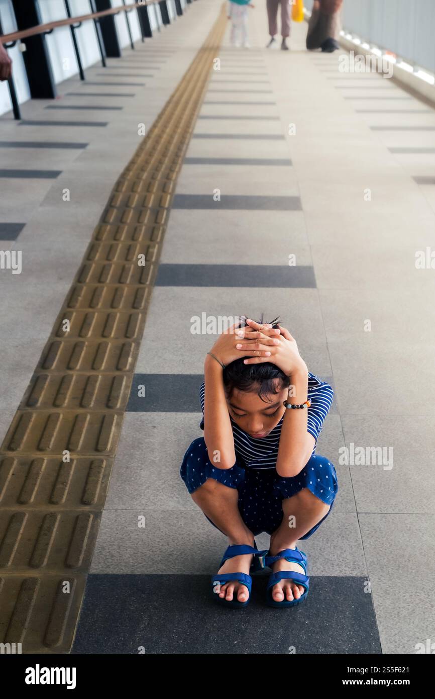 Ragazzo del sud-est asiatico accovacciato con le mani sopra la testa mostrando gesti ed espressioni stanchi al centro di un passaggio pedonale su un ponte di collegamento Foto Stock
