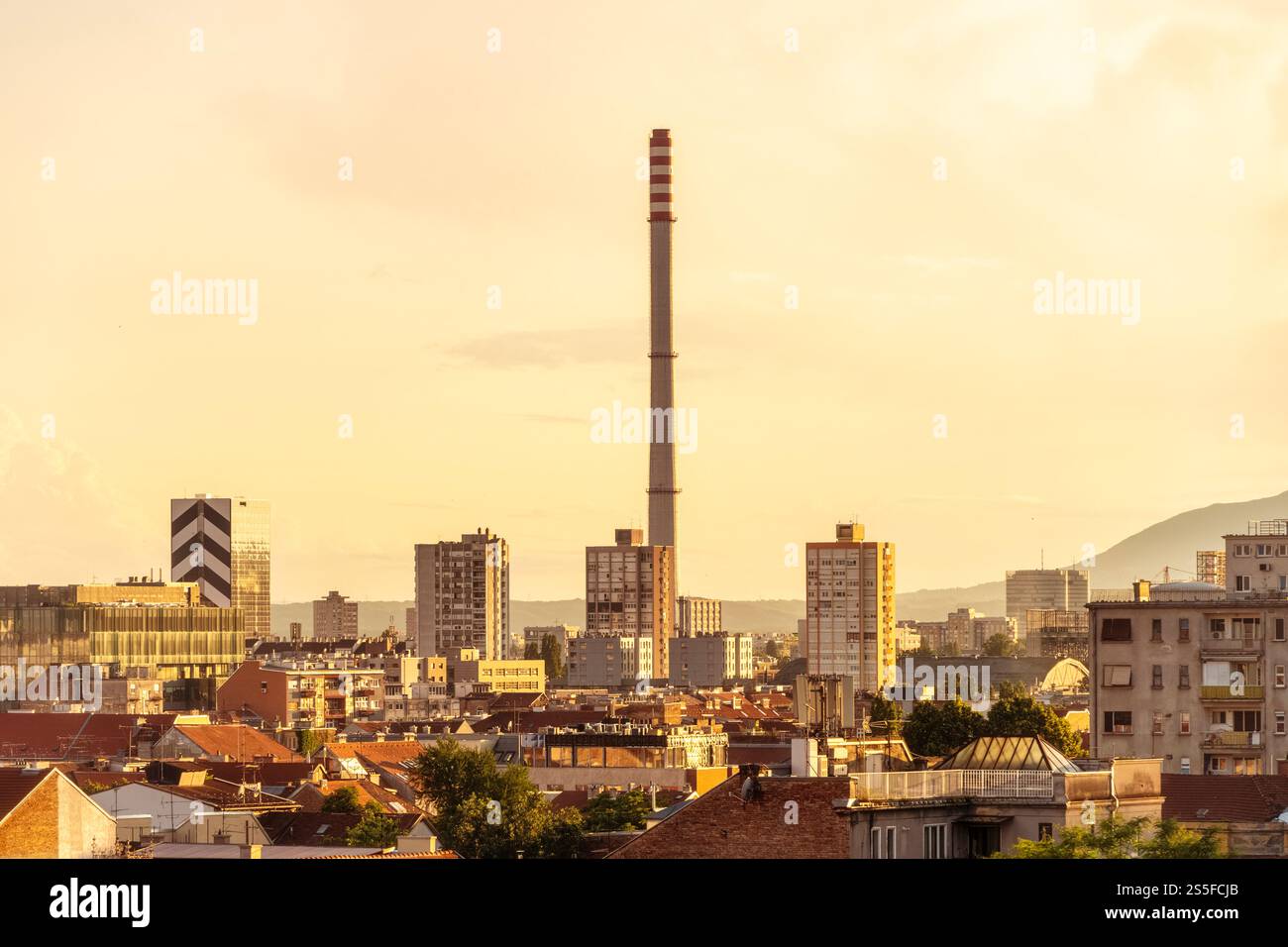 Tramonto sullo skyline della città di Zagabria con un alto camino industriale ed edifici residenziali contro un cielo nuvoloso, Zageb, Croazia Foto Stock