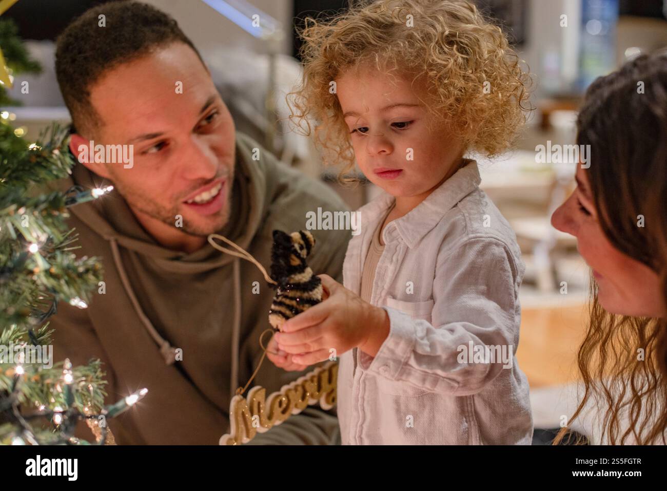 Una famiglia adora decorare un albero di Natale insieme, con un bambino che regge un ornamento "allegro", Ohio, Stati Uniti Foto Stock