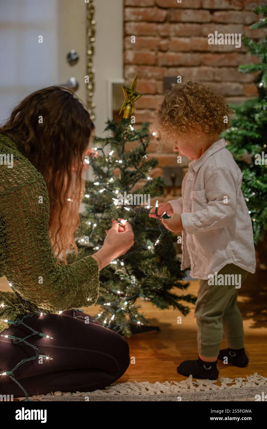 Una donna e un bambino stanno decorando un albero di Natale, con la donna che organizza le luci e il bambino che arriva verso l'albero con fascino, Ohio, Stati Uniti Foto Stock
