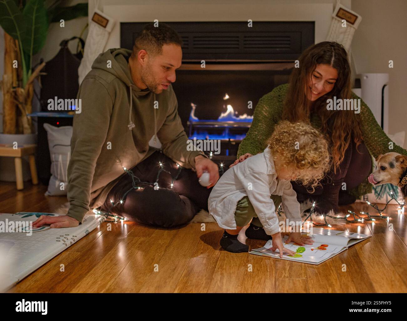 La famiglia si diverte con un bambino e un cane accanto al caminetto, circondato da luci scintillanti, Ohio, USA Foto Stock