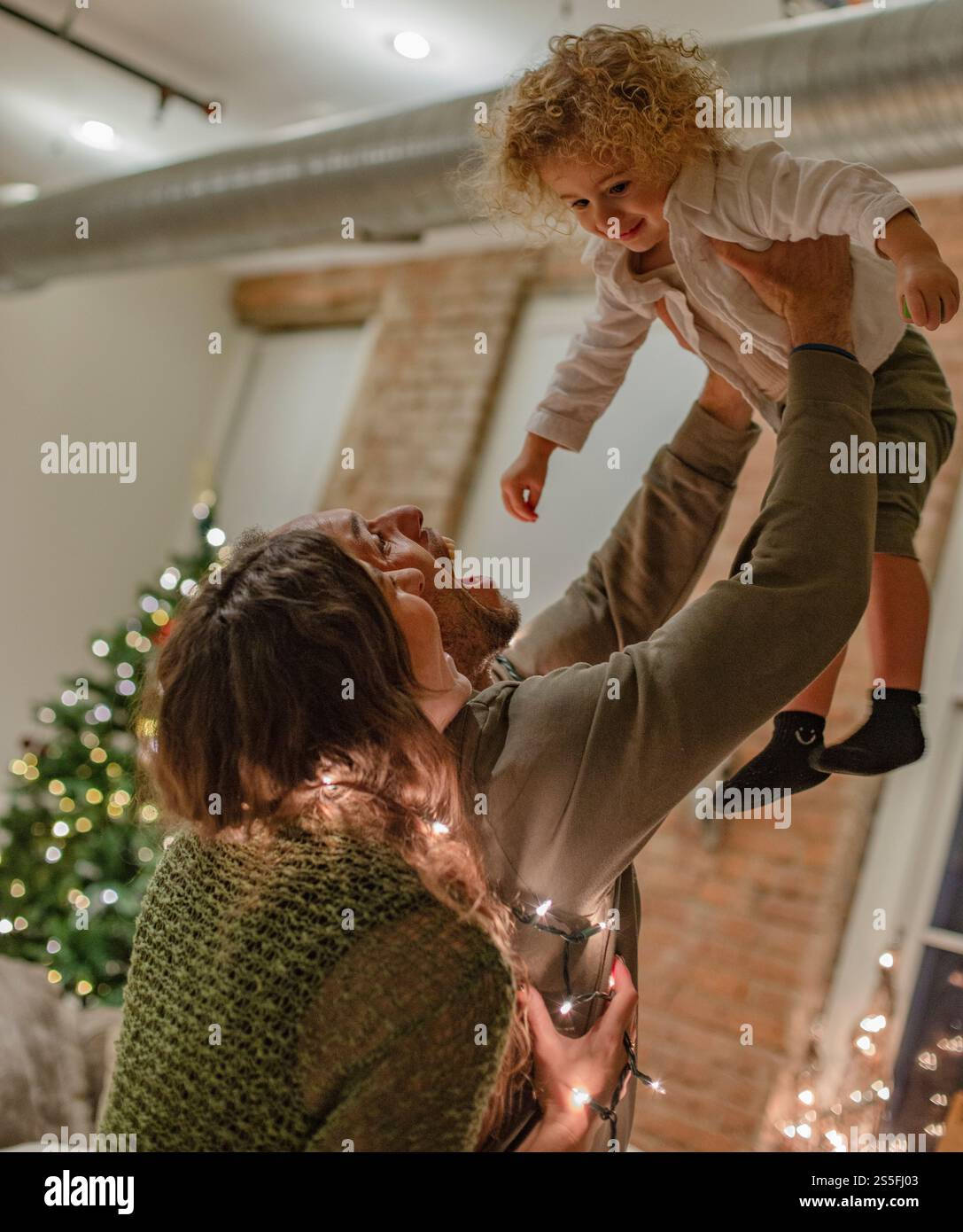 Un padre gioioso solleva il bambino in aria in una stanza accogliente decorata con luci natalizie e un albero, Ohio, USA Foto Stock
