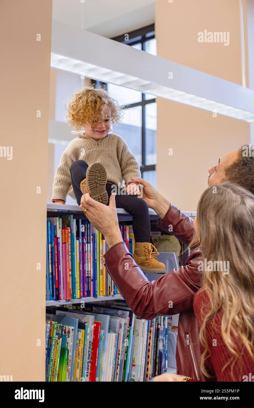 Bambino sorridente che viene sollevato da un adulto in una biblioteca con libri colorati sullo sfondo. Cleveland, Ohio, Stati Uniti Foto Stock