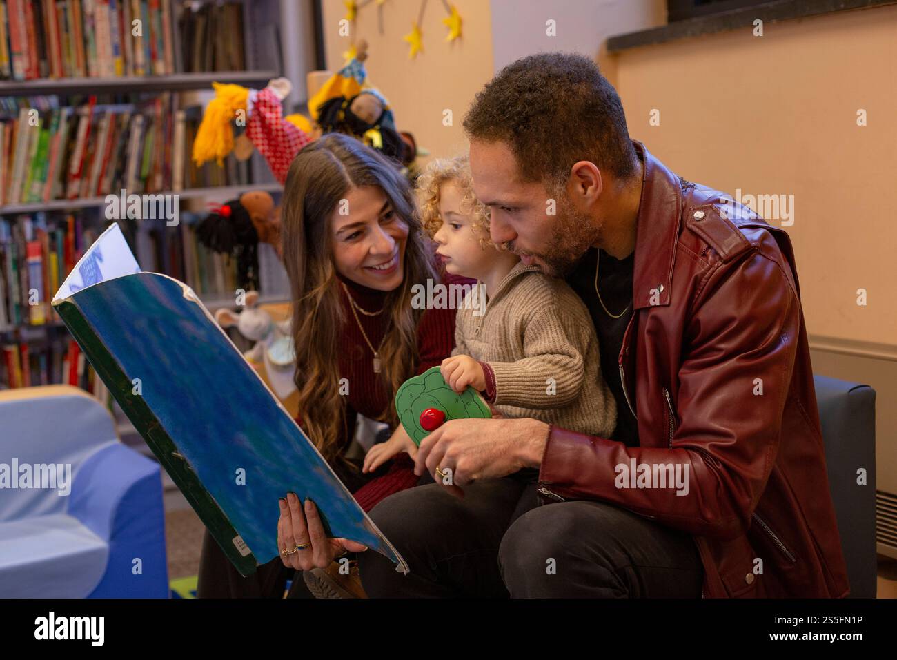 Una famiglia che si diverte con un libro in una biblioteca con colorati giocattoli per bambini sullo sfondo. Cleveland, Ohio, Stati Uniti Foto Stock