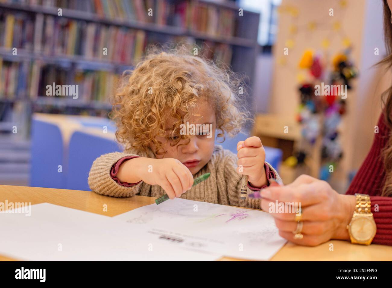 Bambino dai capelli ricci concentrato sul disegno con pastelli a un tavolo in una biblioteca mentre una persona si siede accanto a loro. Cleveland, Ohio, Stati Uniti Foto Stock
