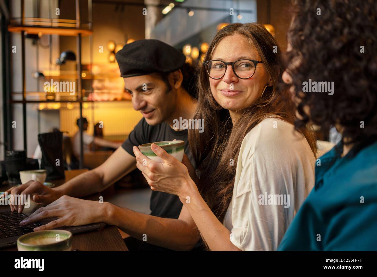 Tre amici o colleghi che condividono un momento in un accogliente bar, con un uomo che digita su un laptop e un'altra donna che si occupa di conversazione, Berlino, Germania Foto Stock