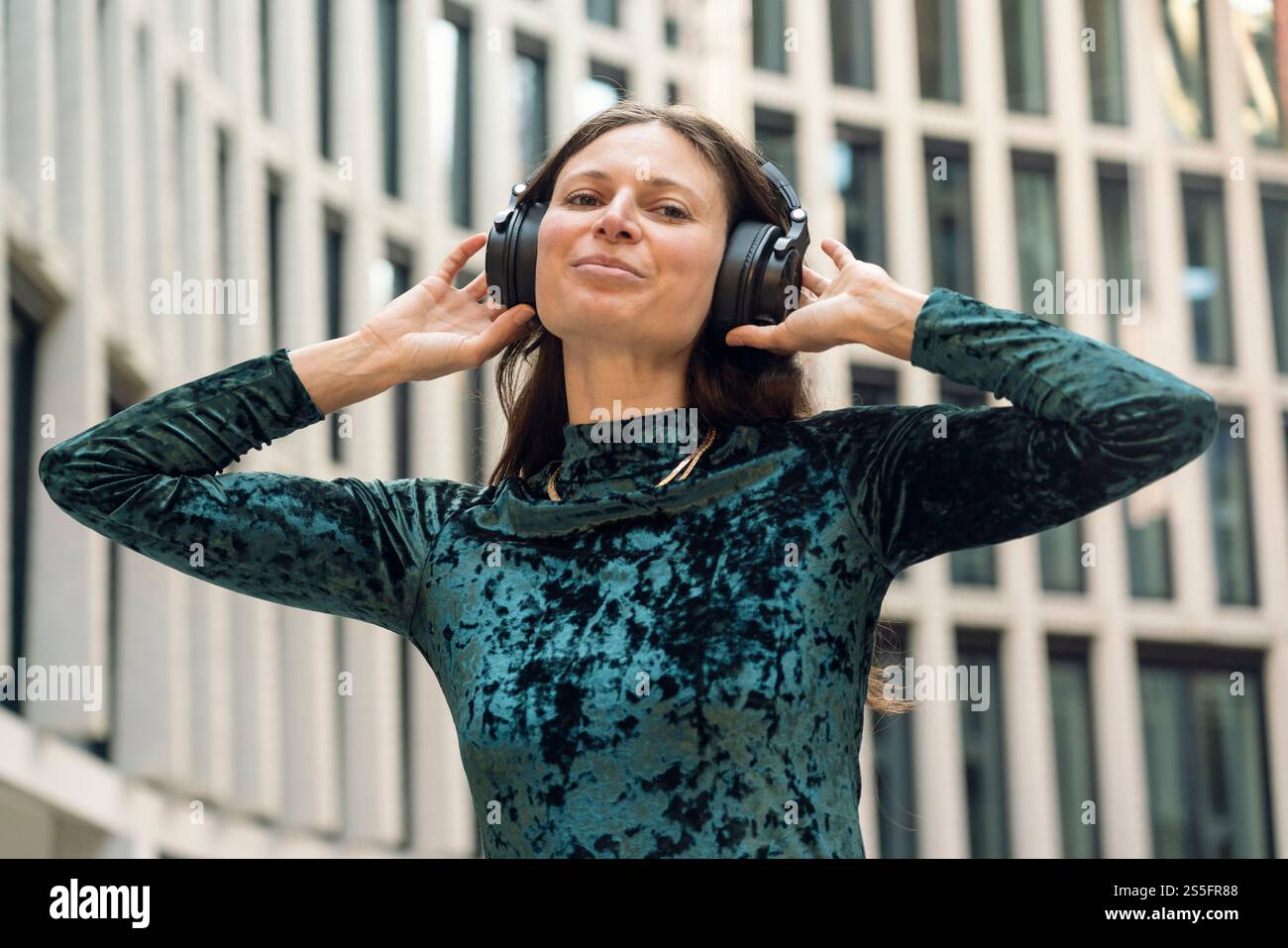 Una donna sorridente con un motivo blu ama ascoltare musica con grandi cuffie nere all'aperto con edifici sullo sfondo, Berlino, Germania Foto Stock
