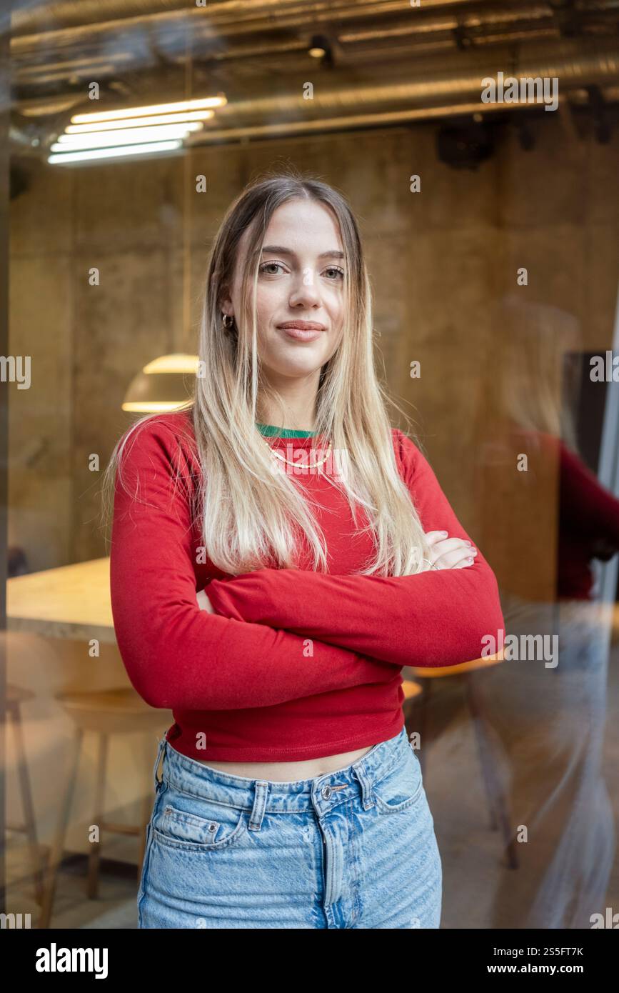 Giovane donna sicura di sé con un top rosso e jeans blu in piedi con le braccia incrociate in un ambiente d'ufficio, Berlino, Germania Foto Stock