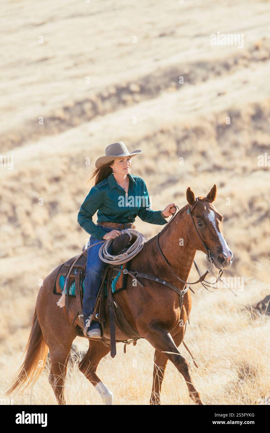 Una donna con un cappello a teglia larga cavalca un cavallo marrone attraverso un paesaggio asciutto e illuminato dal sole, tenendo una corda a spirale e guidando il suo cavallo con competenza, Idaho, USA Foto Stock