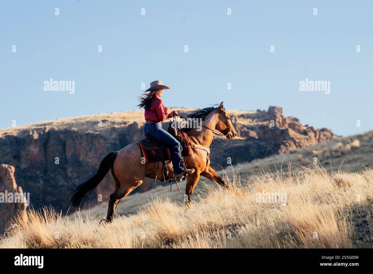 Una donna che indossa un cappello a tesa larga e una maglietta rossa cavalca su una collina erbosa durante una giornata limpida, con un paesaggio roccioso sullo sfondo, Idaho, Stati Uniti Foto Stock