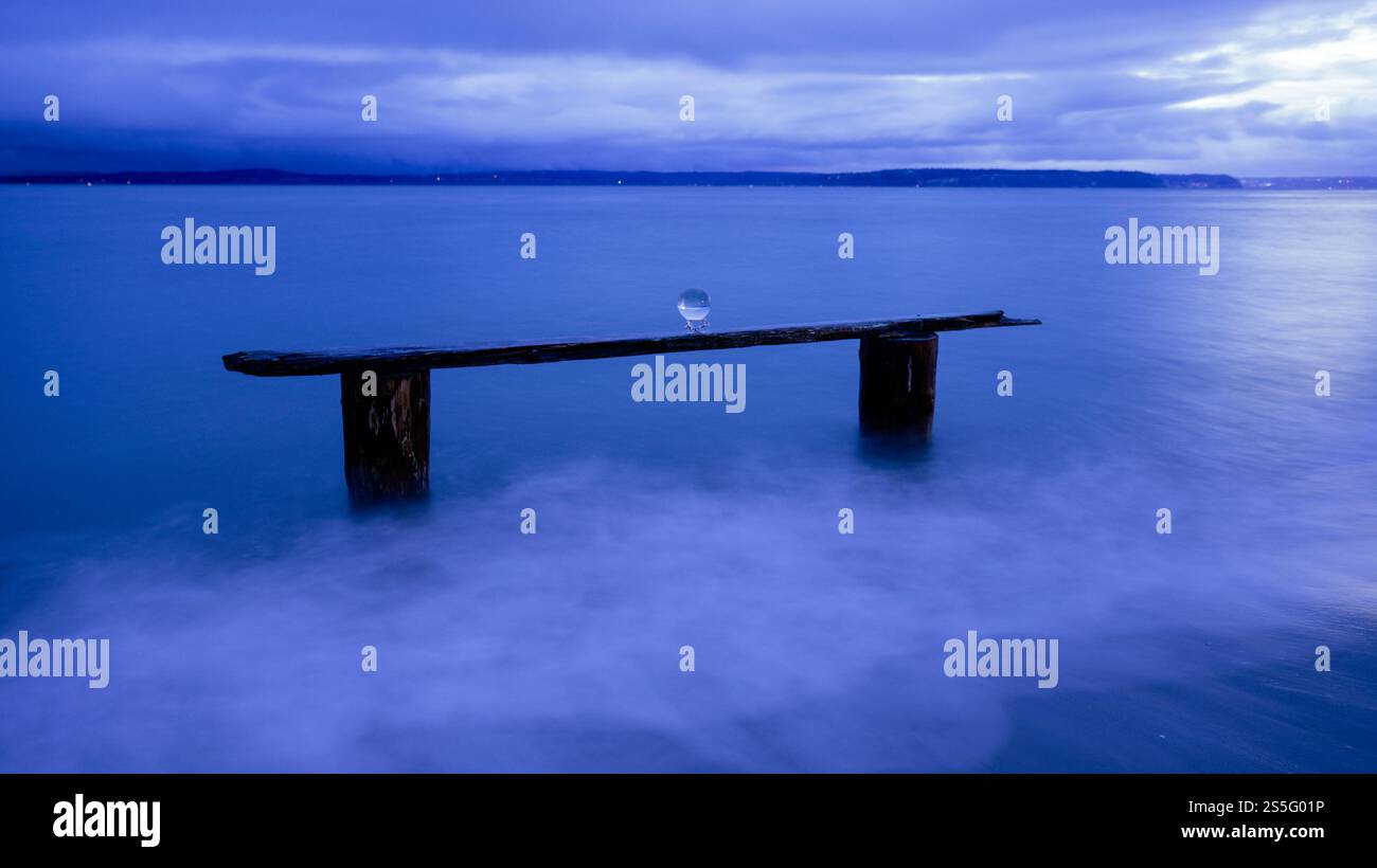 Un tranquillo paesaggio marino al crepuscolo presenta un cielo azzurro e un oceano calmo, con una vecchia trave di legno che sostiene una sfera di cristallo, Norwegian Point, WA, USA Foto Stock