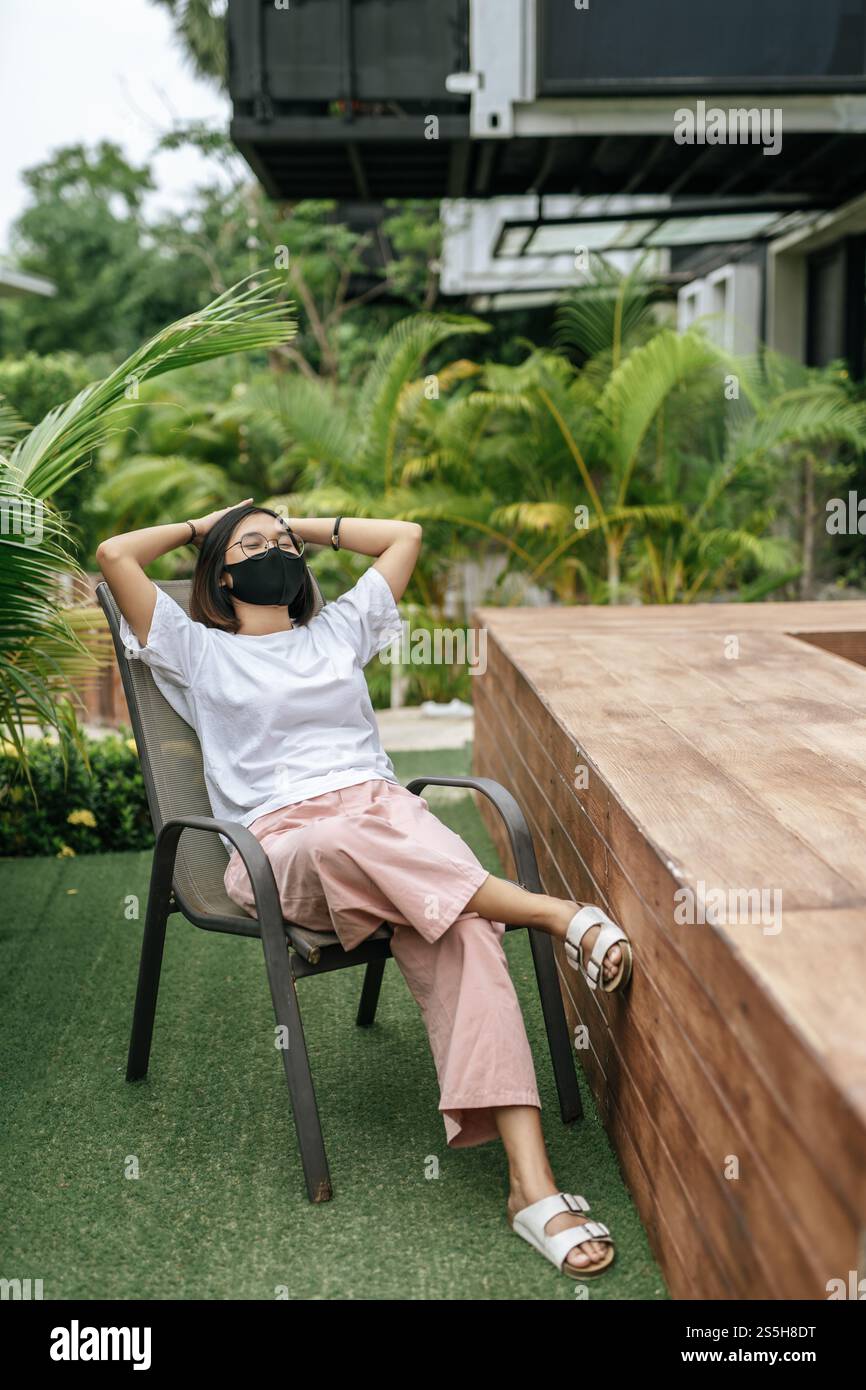Donna che indossa una maschera seduta su una sedia accanto a una piscina in giardino. Foto Stock