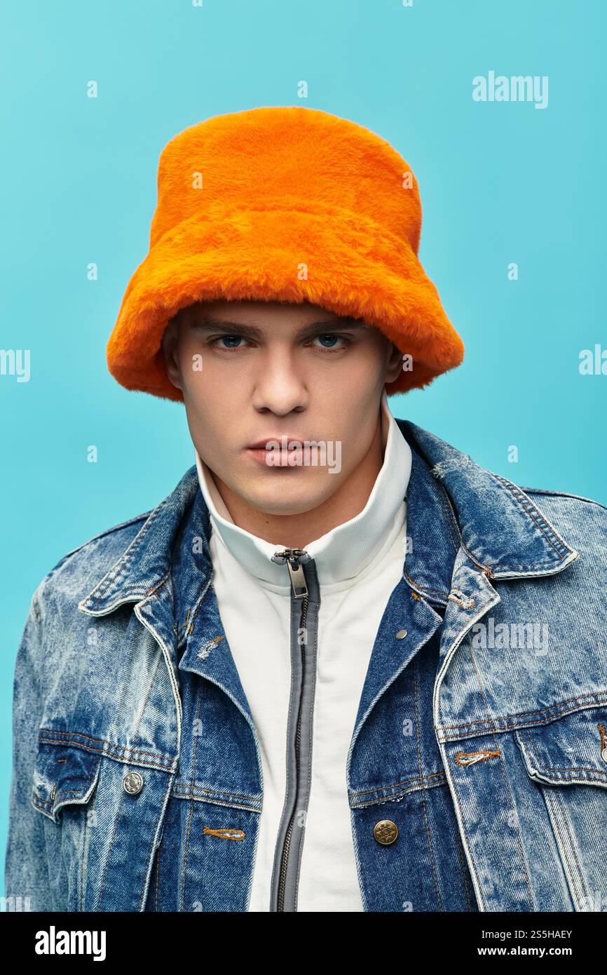 Il bellissimo giovane presenta un elegante cappello arancione, vestito in denim, su uno sfondo blu brillante. Foto Stock