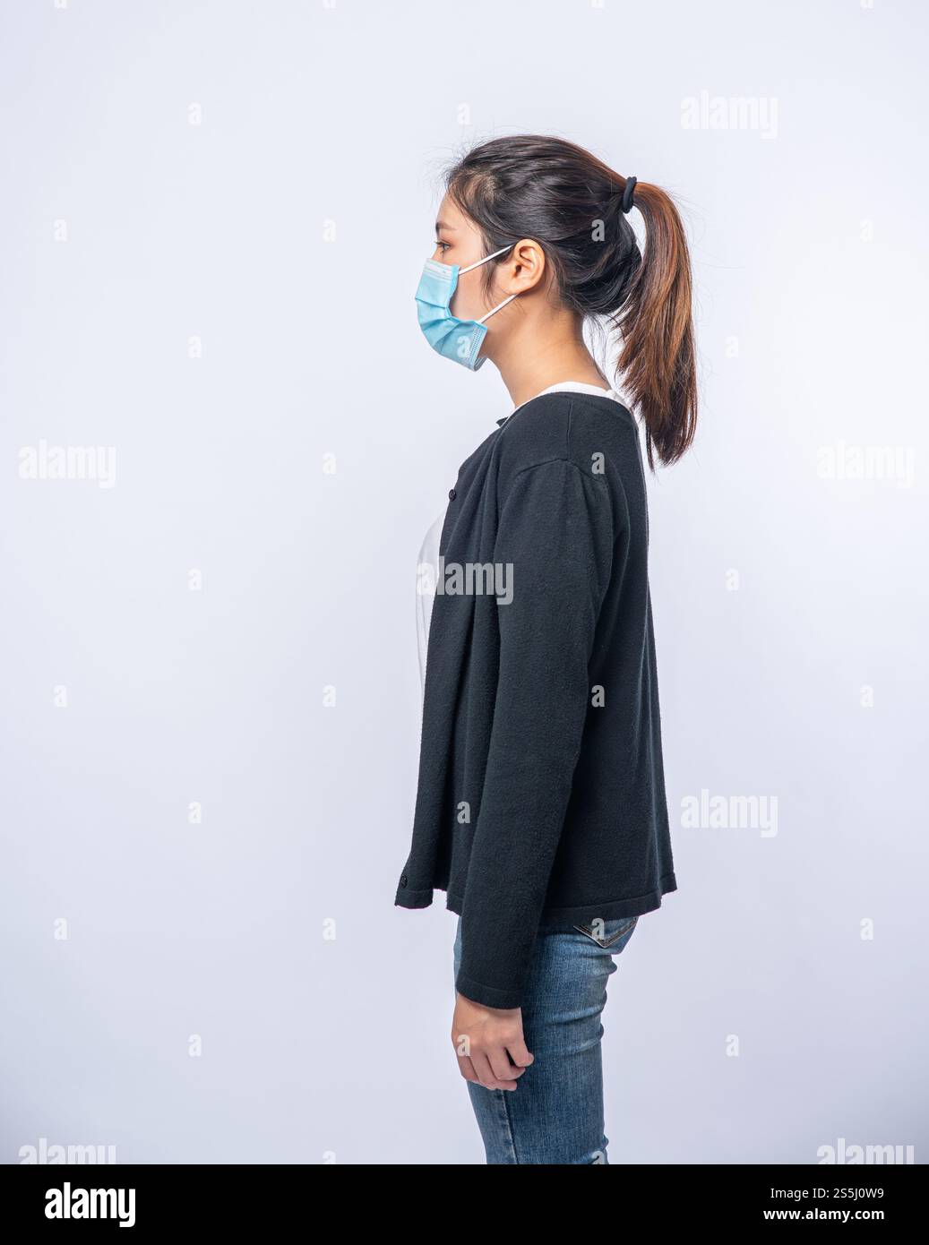 Una donna è malata in piedi e indossa una maschera. Indossa un cappotto nero e un paio di jeans. Foto Stock