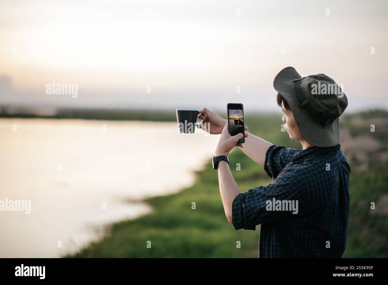 Giovane uomo che indossa un cappello da trekking felice di usare lo smartphone scatta una foto con una tazza di caffè sulla splendida natura sullo sfondo Foto Stock