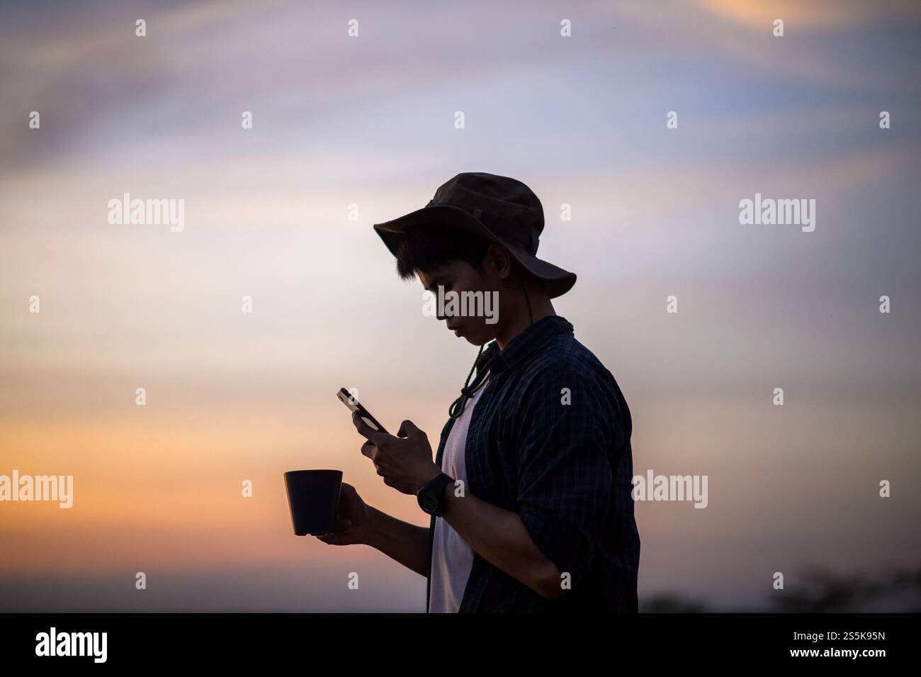 Giovane uomo che indossa un cappello da trekking felice di usare lo smartphone scatta una foto con una tazza di caffè sulla splendida natura sullo sfondo Foto Stock
