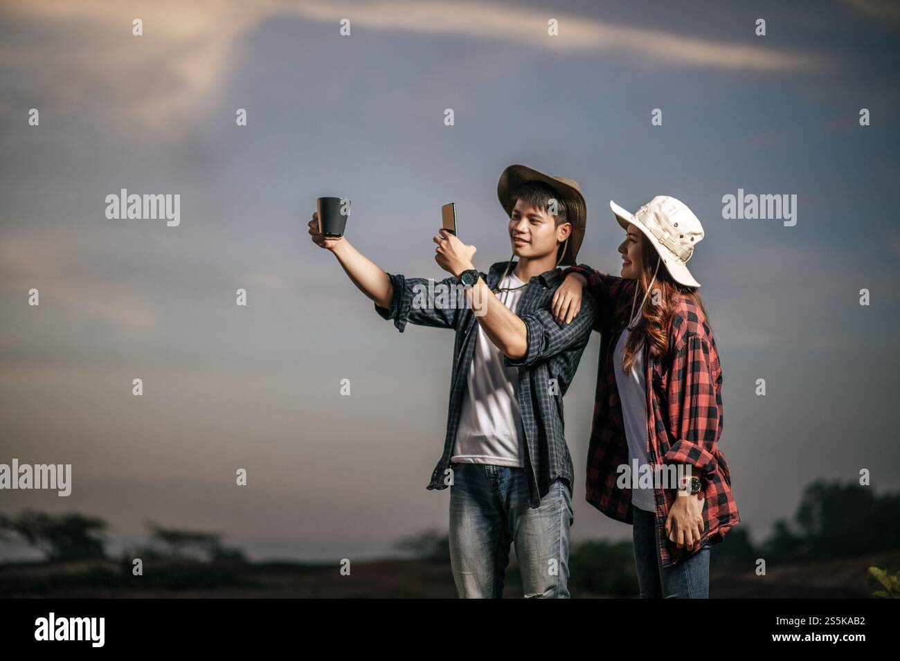 Un giovane che indossa un cappello da trekking felice di usare lo smartphone scatta una foto con una tazza di caffè sullo sfondo, una bella ragazza in piedi Foto Stock