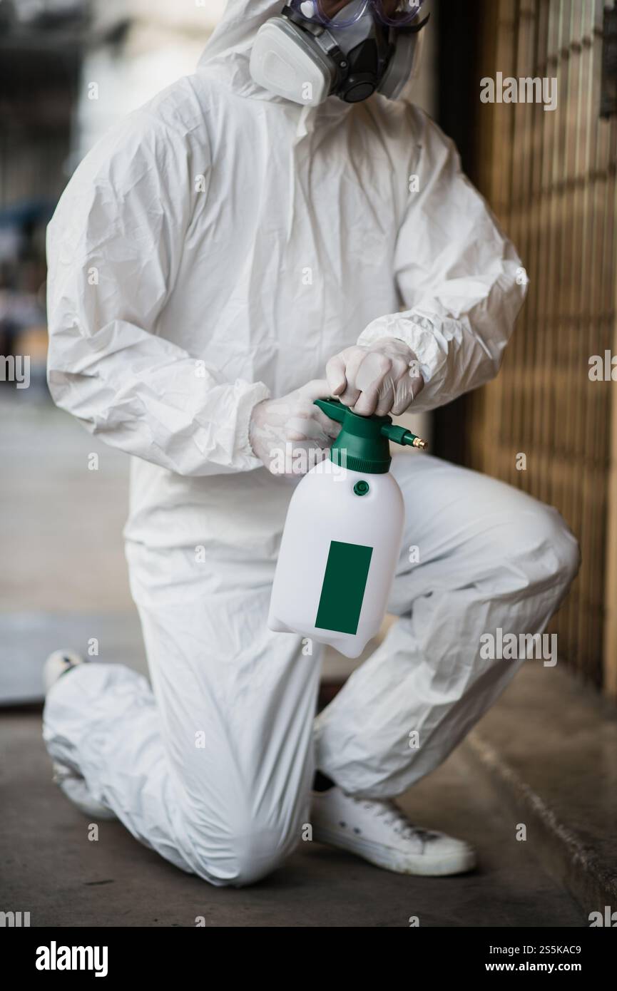 Specialista di disinfezione uomo che indossa i dispositivi di protezione individuale (dpi) tuta, guanti, maschera e occhiali trasparenti, casa di pulizia con bottiglia di Foto Stock