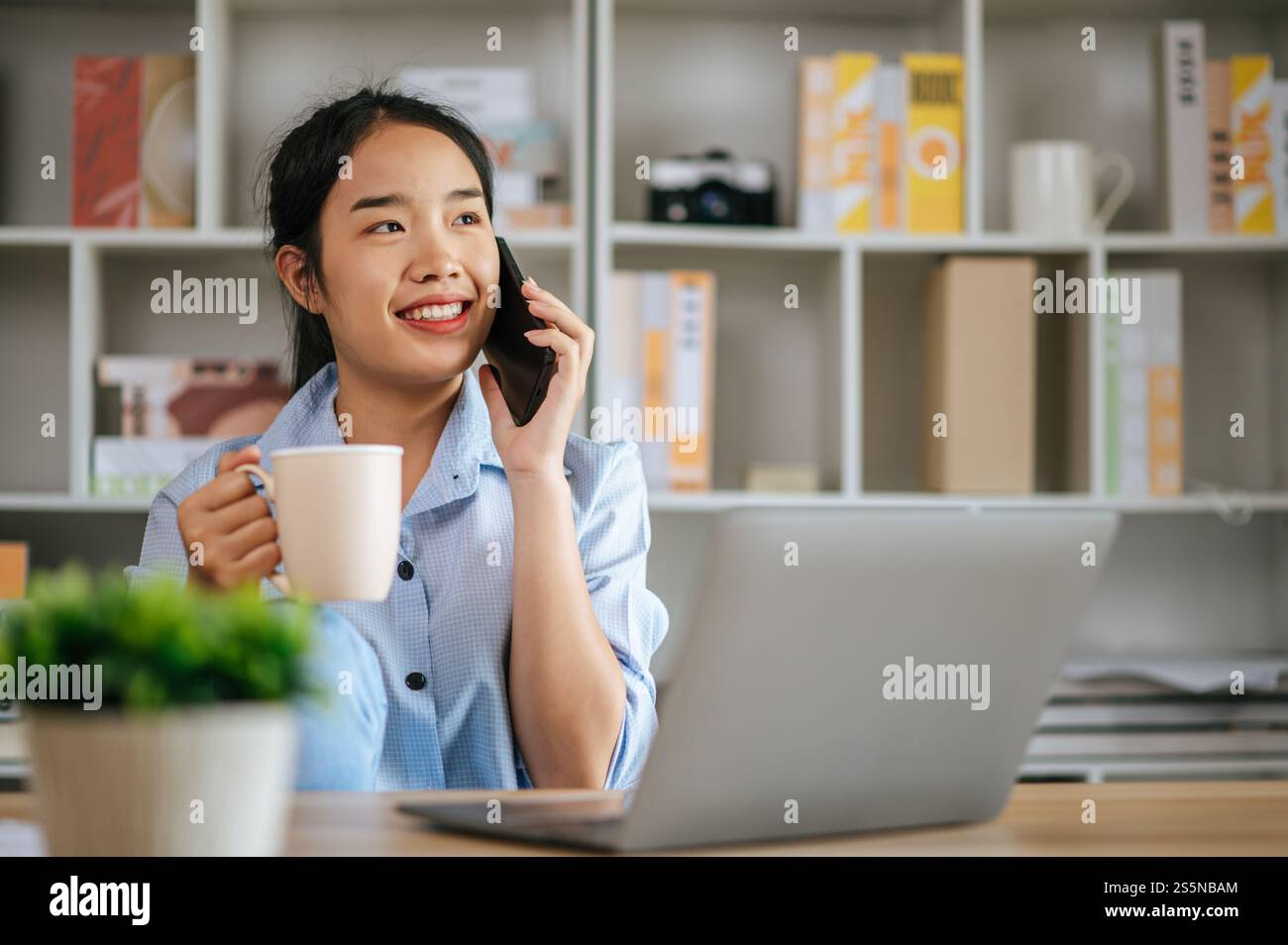 Allegra giovane bella donna che parla con lo smartphone e usa il computer portatile per lavorare o imparare online, tieni la tazza di caffè in mano e sorridi con gioia Foto Stock