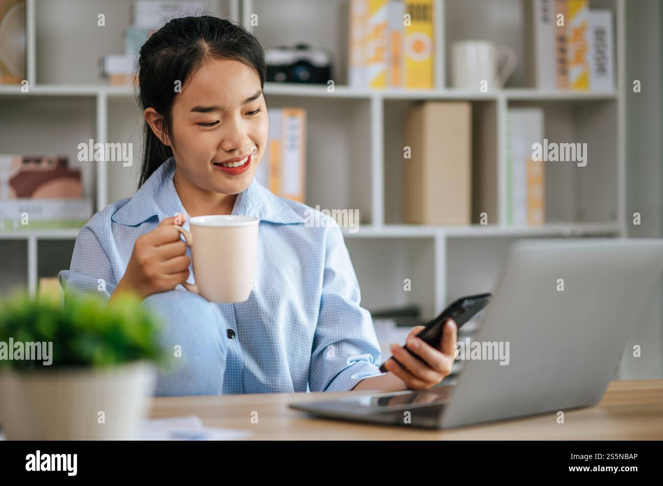 Allegra giovane bella donna usa il computer portatile e lo smartphone per lavorare o imparare online, tieni la tazza di caffè in mano e sorridi con felice durante Foto Stock