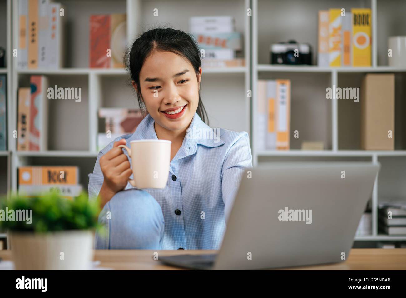 Allegra giovane bella donna seduta e usa il computer portatile e il libro di testo per lavorare o imparare online, tenendo in mano la tazza di caffè e sorridendo con gioia Foto Stock