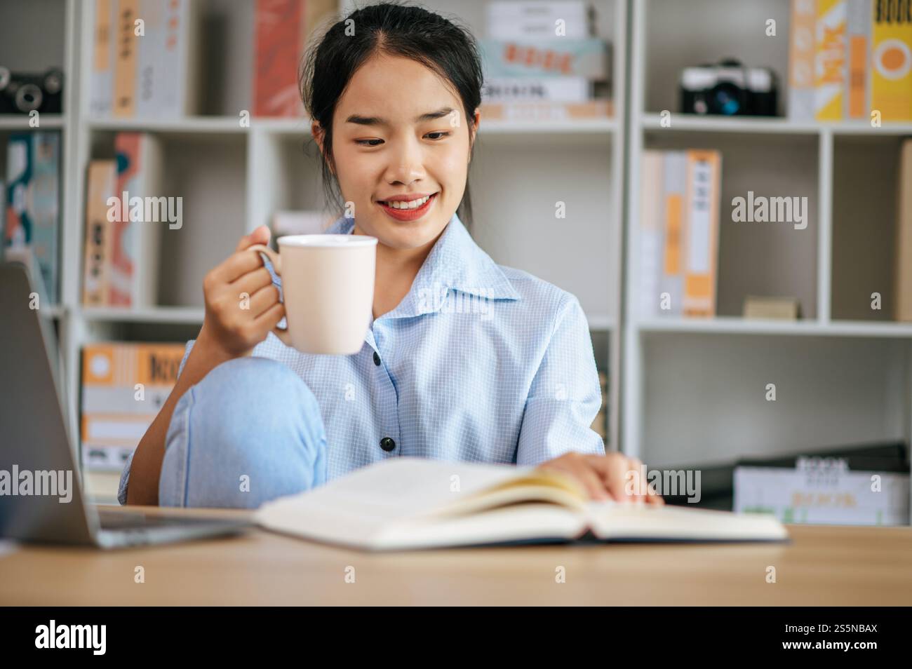 Allegra giovane bella donna seduta e usa il computer portatile e il libro di testo per lavorare o imparare online, tenendo in mano la tazza di caffè e sorridendo con gioia Foto Stock