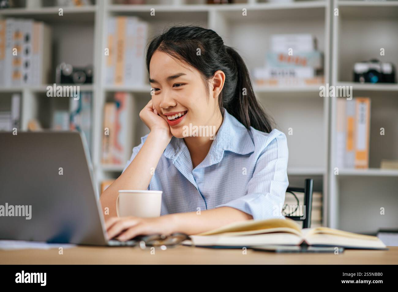 Allegra giovane bella donna seduta e usa il computer portatile e il libro di testo per lavorare o imparare online, tenendo in mano la tazza di caffè e sorridendo con gioia Foto Stock