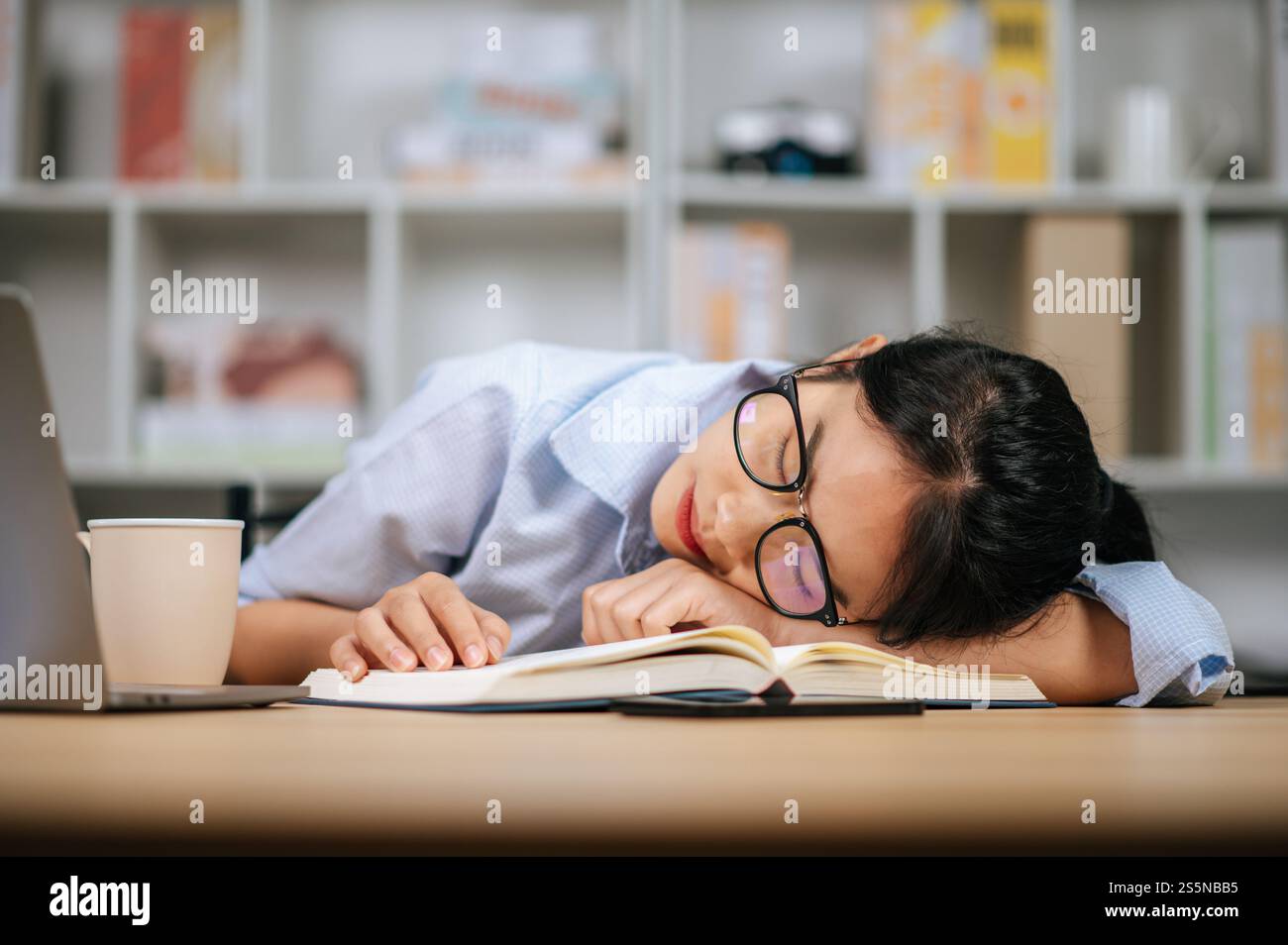 Giovane donna asiatica stanca con gli occhiali da vista che dorme sulla scrivania con un portatile e un libro di testo a casa Foto Stock
