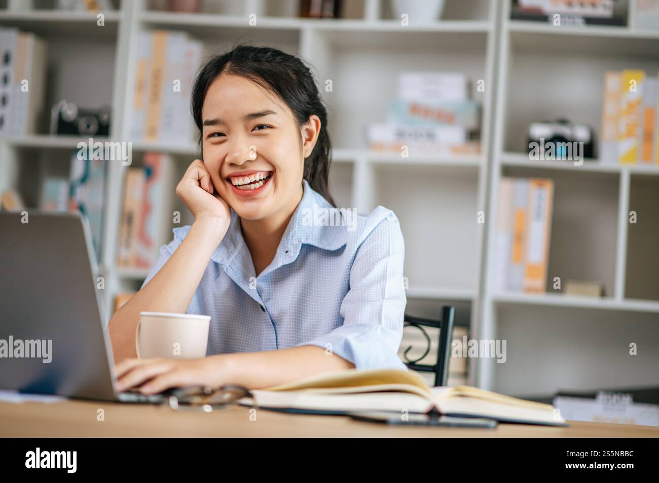 Allegra giovane bella donna seduta e usa il computer portatile e il libro di testo per lavorare o imparare online, tenendo in mano la tazza di caffè e sorridendo con gioia Foto Stock