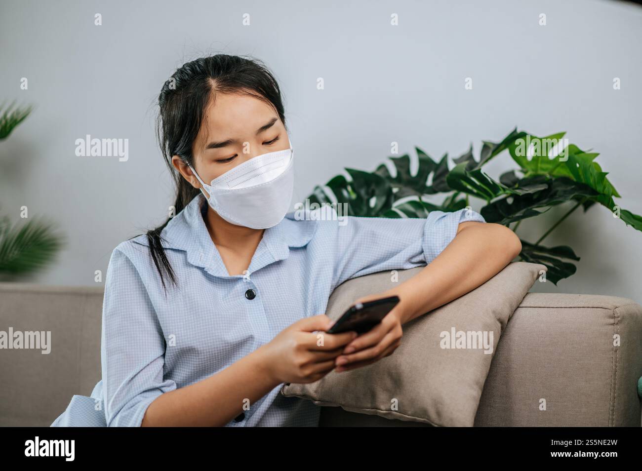 Smiley bella adolescente in maschera seduta sul divano e utilizzare lo smartphone per videochiamare o selfie, durante la quarantena covid-19 auto isolamento a casa Foto Stock
