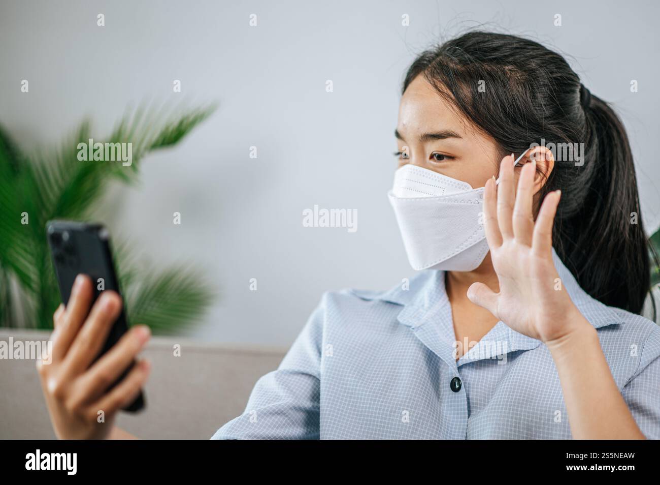 Smiley bella adolescente in maschera seduta sul divano e utilizzare lo smartphone per videochiamare o selfie, durante la quarantena covid-19 auto isolamento a casa Foto Stock