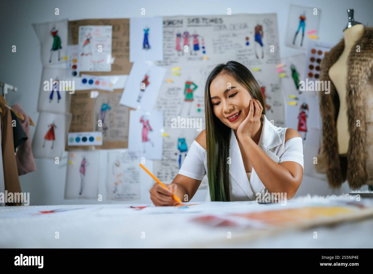 Giovane stilista asiatica donna disegno moda schizzo in officina. Bellissimi vestiti disegnano foto a bordo dietro di lei Foto Stock