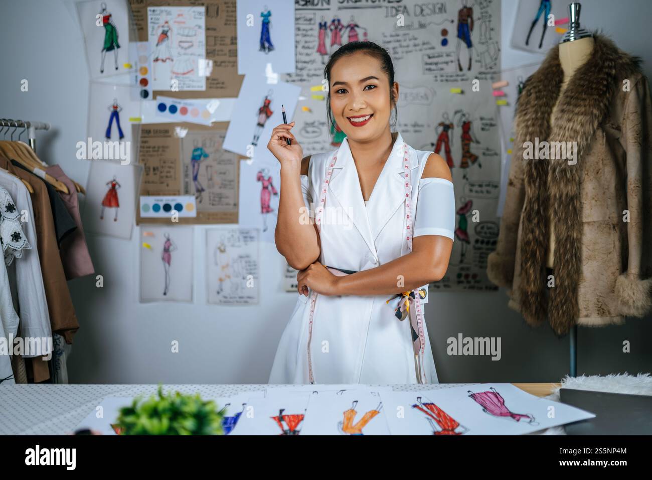 Ritratto giovane stilista asiatica donna disegno disegno moda schizzo in officina. Bellissimi vestiti disegnano foto a bordo dietro di lei Foto Stock