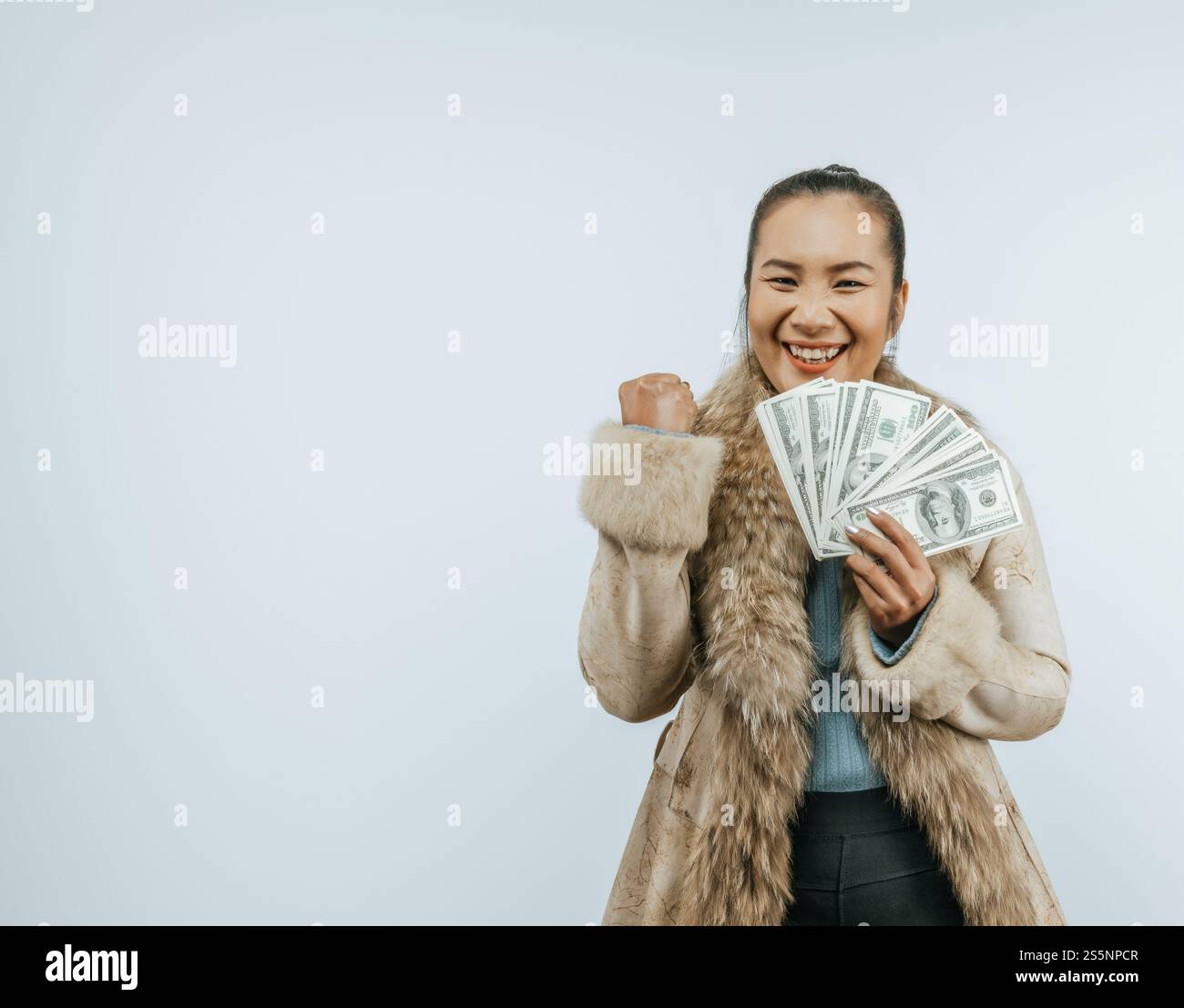 Ritratto di una donna asiatica di successo con un cappotto di pelliccia che tiene in mano i soldi, stringe il pugno, sorride e guarda la fotocamera, in piedi sullo sfondo Foto Stock