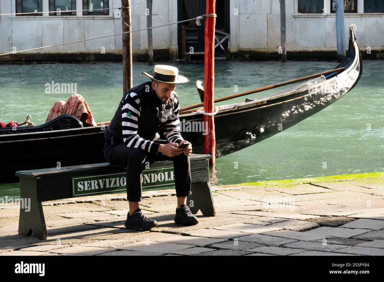 Un gondoliere si siede con il suo telefono vicino a un canale a Venezia, in Italia. Foto Stock