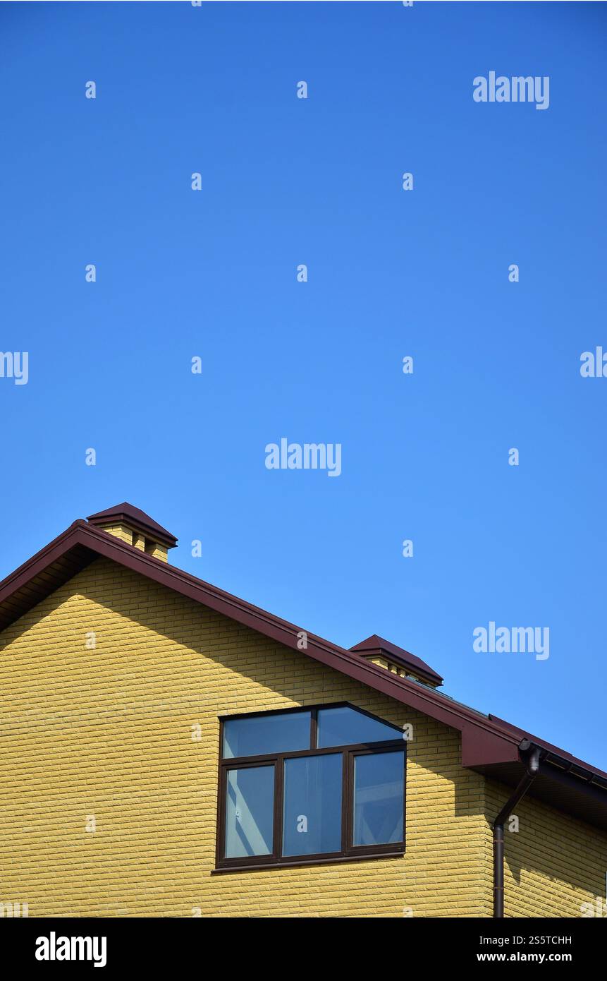 La cima di un edificio residenziale di mattoni gialli con un tetto marrone e finestre di plastica contro un cielo blu chiaro. Il concetto di copertura o esterno Foto Stock
