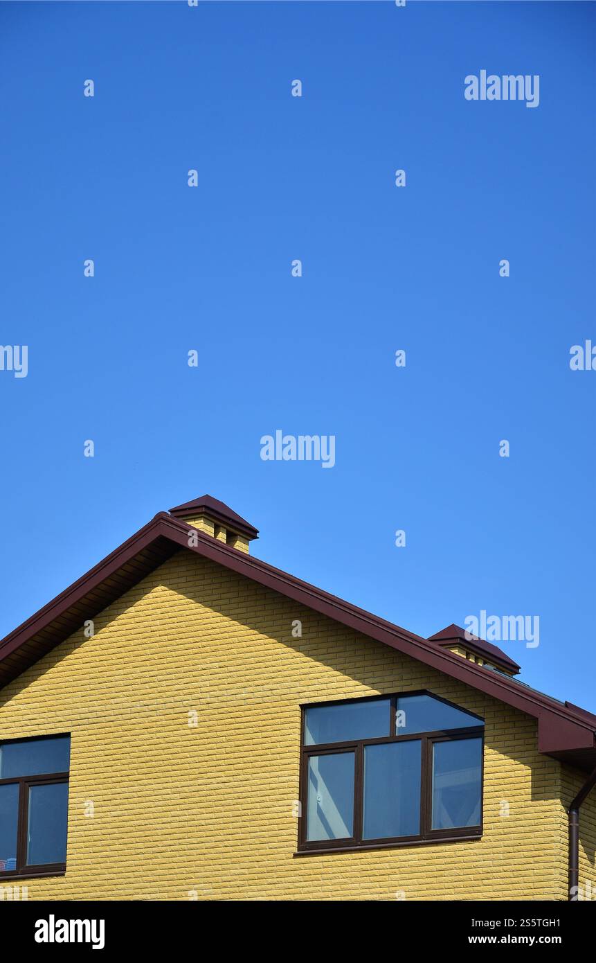 La cima di un edificio residenziale di mattoni gialli con un tetto marrone e finestre di plastica contro un cielo blu chiaro. Il concetto di copertura o esterno Foto Stock